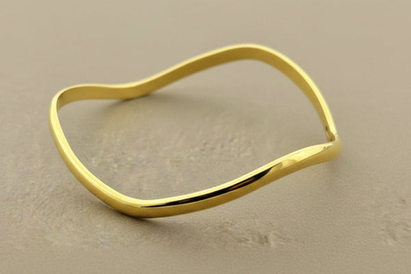 A polished gold-toned metal bangle bracelet with an organic, wavy outline, displayed on a textured gray background.