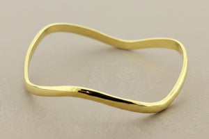 A polished gold-toned metal bangle bracelet with a wavy, organic silhouette, displayed on a textured dark gray background.