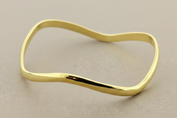 A polished gold-toned metal bangle bracelet with a wavy, organic silhouette, displayed on a textured dark gray background.