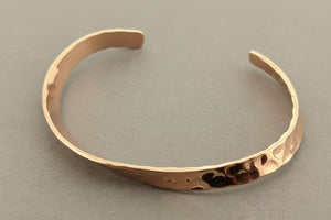 A hammered open cuff bracelet in a warm coppery metallic finish, photographed on a dark gray textured background.