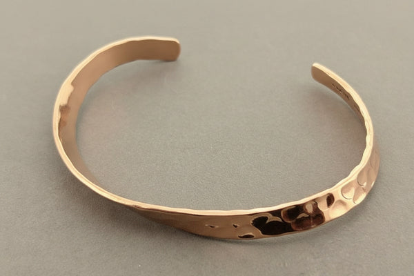 A hammered open cuff bracelet in a warm coppery metallic finish, photographed on a dark gray textured background.
