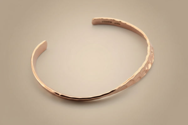 A hammered open cuff bracelet in a warm rose gold metal, displayed on a dark gray textured background.