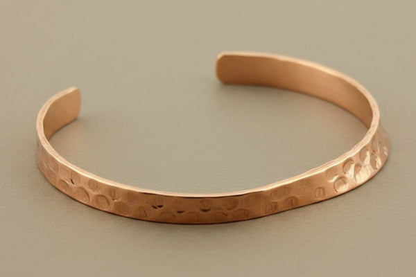 A hammered copper cuff bracelet with a textured, organic finish, displayed on a dark gray background.