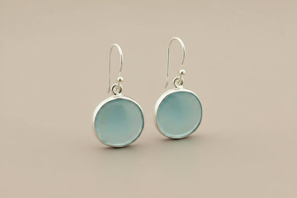 A pair of silver drop earrings with round, light blue sea glass disc pendants, displayed on a dark gray textured background.