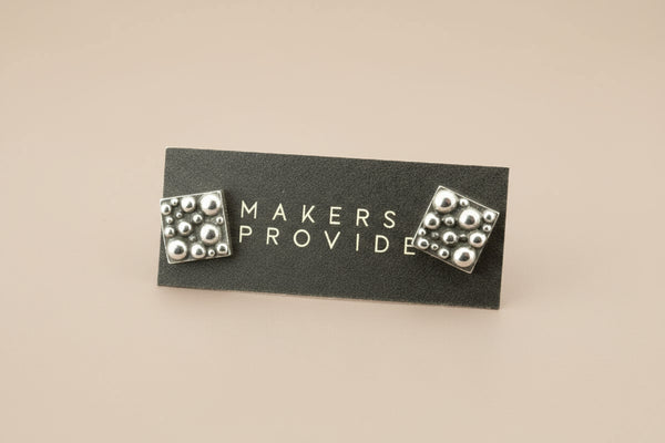 Pair of silver stud earrings on a black card with 'MAKERS PROVIDE' text.
