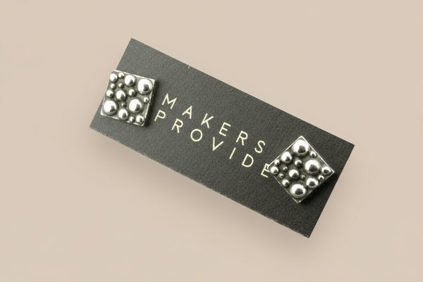 Silver stud earrings on a 'MAKERS PROVIDE' card against a gray background