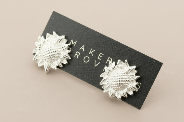 Silver sunburst earrings on a black card with ' branding against a gray background