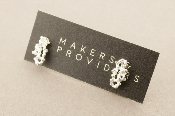 Silver earrings on a 'Makers Provides' card against a gray background