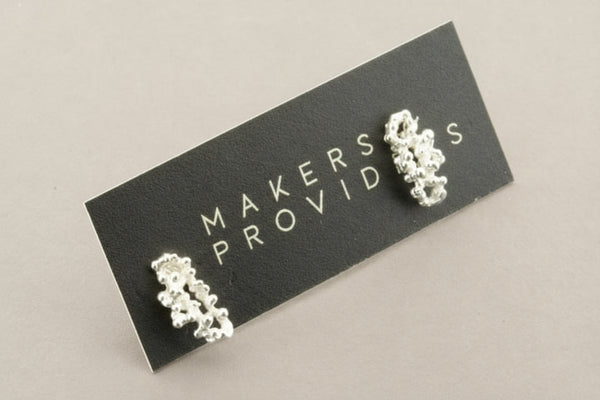 Silver earrings on a 'Makers Provide' card against a gray background