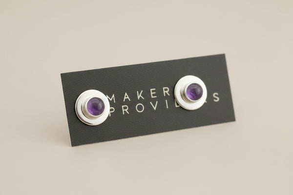 Pair of silver earrings with purple stones on a 'Maker Provides' card against a gray background