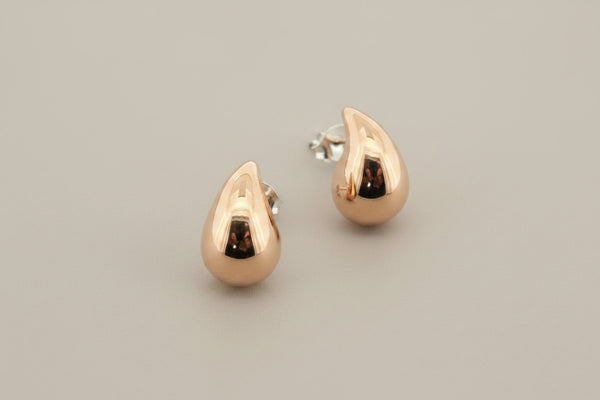 A pair of polished rose gold teardrop-shaped stud earrings with silver posts, displayed on a textured dark gray background.