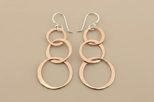 A pair of handmade dangle earrings featuring three interlocking polished rose gold rings on each earring, suspended from silver fishhook ear wires, displayed on a dark gray textured background.