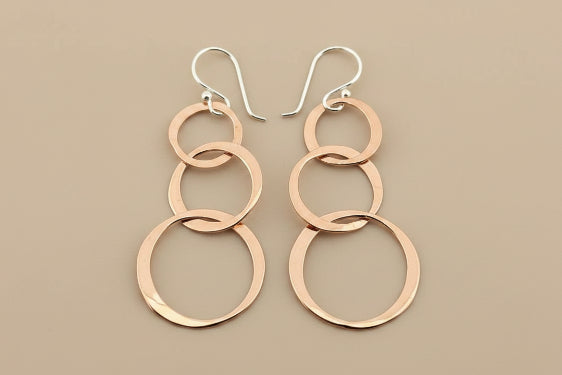 A pair of handmade dangle earrings featuring three interlocking polished rose gold rings on each earring, suspended from silver fishhook ear wires, displayed on a dark gray textured background.