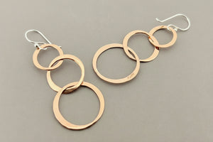 A pair of dangle earrings made of overlapping brushed copper-colored metal rings with silver fishhook ear wires, displayed on a dark gray textured background.