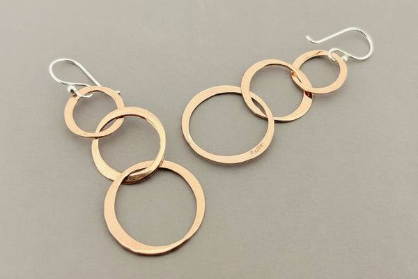 A pair of dangle earrings made of overlapping brushed copper-colored metal rings with silver fishhook ear wires, displayed on a dark gray textured background.