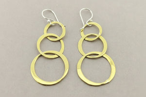 A pair of gold dangle earrings with three stacked interlocking circular rings on each earring, suspended from silver fishhook ear wires, displayed on a dark gray textured background.