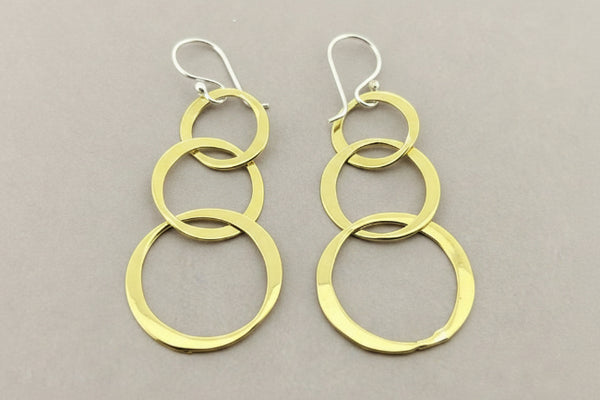 A pair of gold dangle earrings with three stacked interlocking circular rings on each earring, suspended from silver fishhook ear wires, displayed on a dark gray textured background.