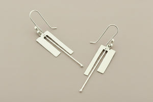 A pair of modern geometric silver drop earrings with rectangular and linear metal elements, displayed on a dark gray textured background.