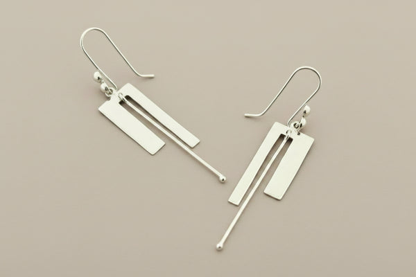 A pair of modern geometric silver drop earrings with rectangular and linear metal elements, displayed on a dark gray textured background.