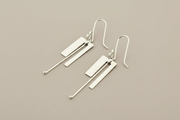A pair of modern silver drop earrings with geometric rectangular and linear elements, displayed on a dark gray textured background.
