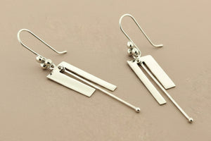 A pair of modern silver dangle earrings with geometric rectangular bars and thin wire hooks, displayed on a dark gray textured background.