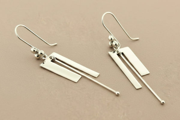 A pair of modern silver dangle earrings with geometric rectangular bars and thin wire hooks, displayed on a dark gray textured background.