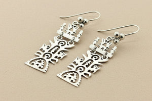 A pair of silver dangling earrings with intricate cutout Celtic knotwork and tree-like patterns, displayed on a dark gray textured background.