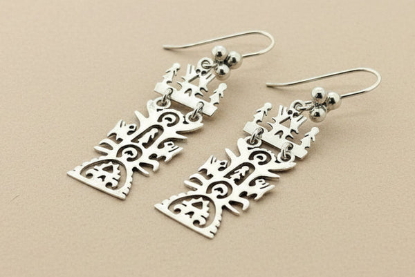 A pair of silver dangling earrings with intricate cutout Celtic knotwork and tree-like patterns, displayed on a dark gray textured background.
