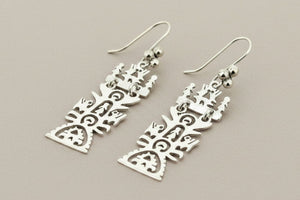 A pair of silver dangle earrings with intricate cutout tribal or abstract geometric patterns, displayed on a dark gray textured background.