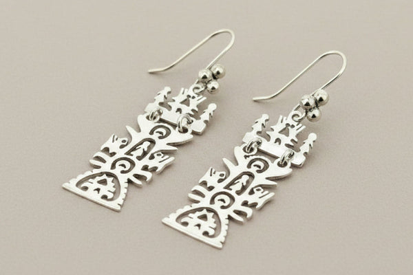 A pair of silver dangle earrings with intricate cutout tribal or abstract geometric patterns, displayed on a dark gray textured background.