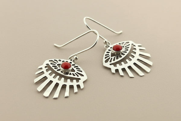 A pair of silver-toned dangle earrings with a fan or sunburst cutout design and a small round red gemstone at the center, shown on a dark gray background.