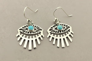 A pair of silver-toned dangle earrings with a fan or sunburst cutout design and a small round turquoise stone at the center, shown on a dark gray background.