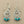 Load image into Gallery viewer, A pair of silver-toned dangle earrings with intricate cutout sunburst designs and three small turquoise beads at the bottom, displayed on a dark gray textured surface.

