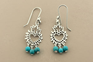 A pair of silver-toned dangle earrings with intricate cutout sunburst designs and three small turquoise beads at the bottom, displayed on a dark gray textured surface.