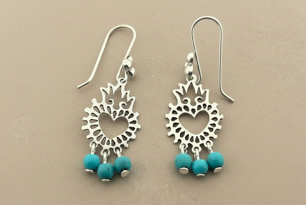A pair of silver-toned dangle earrings with intricate cutout sunburst designs and three small turquoise beads at the bottom, displayed on a dark gray textured surface.