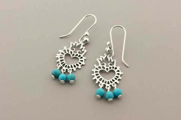 A pair of silver dangle earrings with heart-shaped filigree centers and three small turquoise beads hanging from each earring, displayed on a dark gray textured surface.