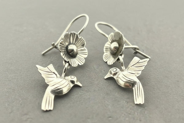 A pair of sterling silver dangle earrings featuring a flower and a hummingbird, displayed on a dark gray textured background.