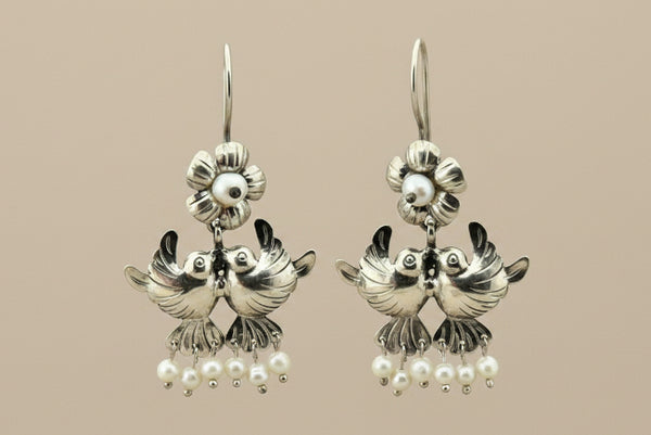 A pair of silver-toned double bird dangle earrings with small white pearl-like beads at the bottom, displayed on a dark gray background.