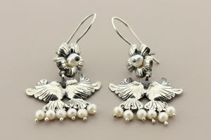 A pair of silver-toned dangling earrings with bird motifs and small pearl-like bead drops, photographed on a dark gray background.