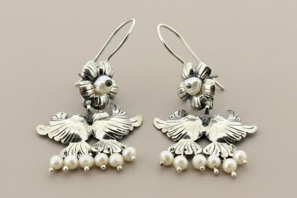A pair of silver-toned dangling earrings with bird motifs and small pearl-like bead drops, photographed on a dark gray background.
