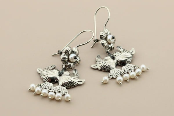 A pair of silver-toned dangle earrings featuring detailed bird motifs with small pearl bead clusters, displayed on a dark gray textured background.