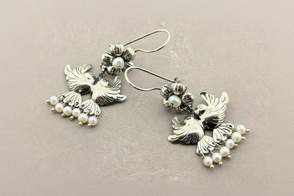 A pair of silver-toned dangle earrings featuring bird motifs with small white pearl bead clusters, displayed on a dark gray textured background.