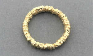 A textured gold band ring with a heavily hammered, organic, and ridged surface, shown on a neutral gray background.