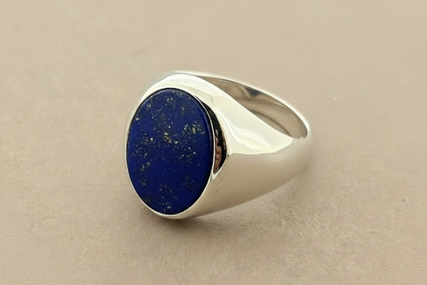 A silver signet ring featuring a round, deep blue lapis lazuli stone with visible golden flecks, set against a dark gray textured background.