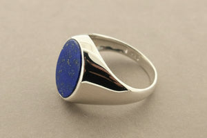 A silver signet ring featuring an oval-cut deep blue lapis lazuli stone, displayed on a textured gray surface.