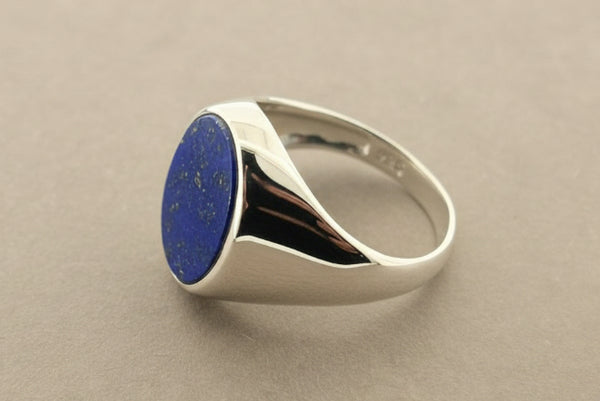 A silver signet ring featuring an oval-cut deep blue lapis lazuli stone, displayed on a textured gray surface.