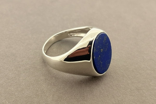 A silver signet ring with a round, polished blue lapis lazuli stone, displayed on a textured dark gray surface.