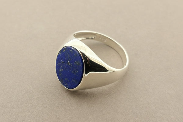 A sterling silver signet ring featuring a round, deep blue lapis lazuli stone, displayed on a textured gray background.
