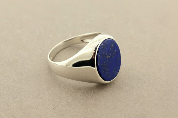 A silver signet ring with a round, deep blue lapis lazuli stone, photographed on a textured gray surface.