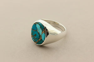 A silver signet ring featuring a round turquoise stone with brown matrix veining, displayed on a dark gray textured surface.